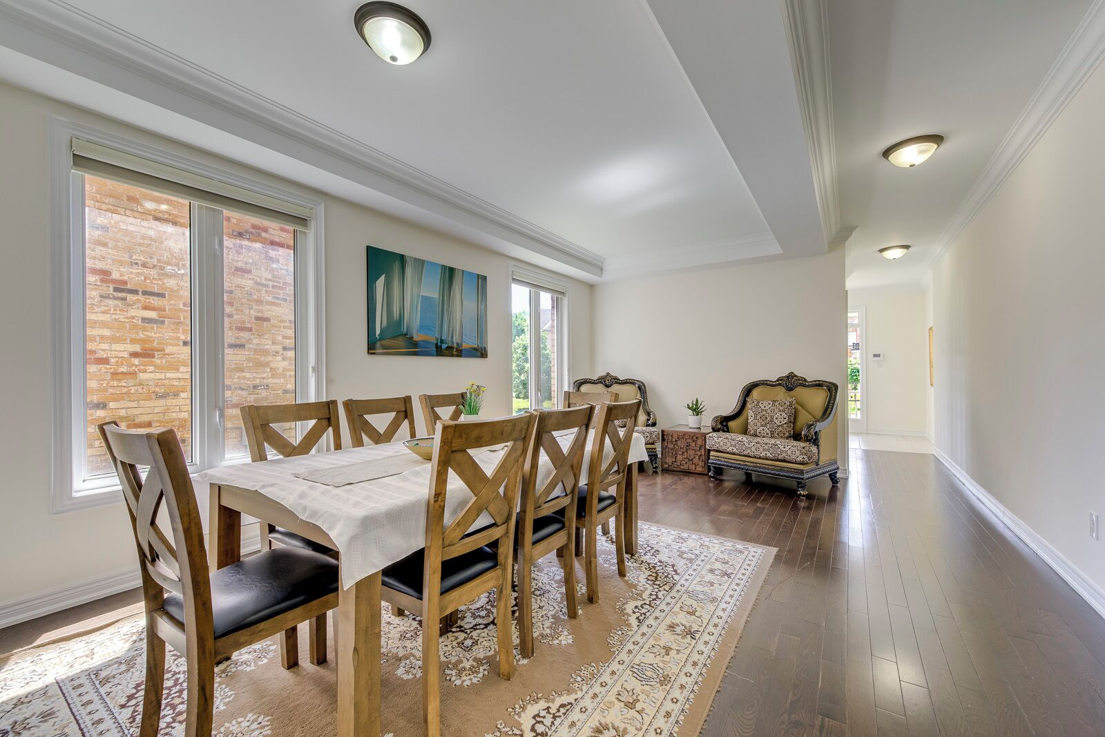 A dining area with a wooden table and chairs, a rug, and a view of a brick wall through large windows.