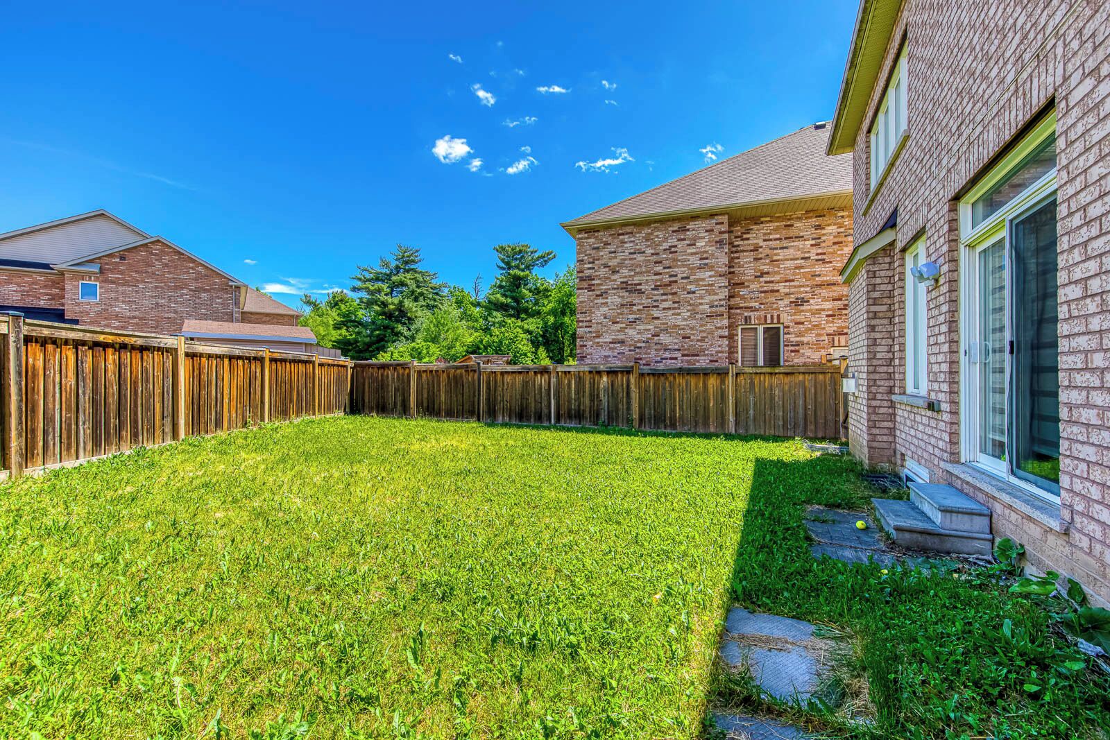 A backyard with a wooden fence, a grassy lawn, and a brick building.