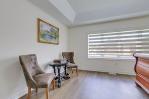 A room with two upholstered chairs, a small round table with a chess set, a painting on the wall, and a window with blinds.