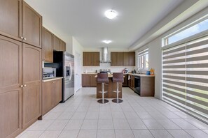 A modern kitchen with wooden cabinets, a stainless steel refrigerator, a central island with bar stools, and a large window with blinds.