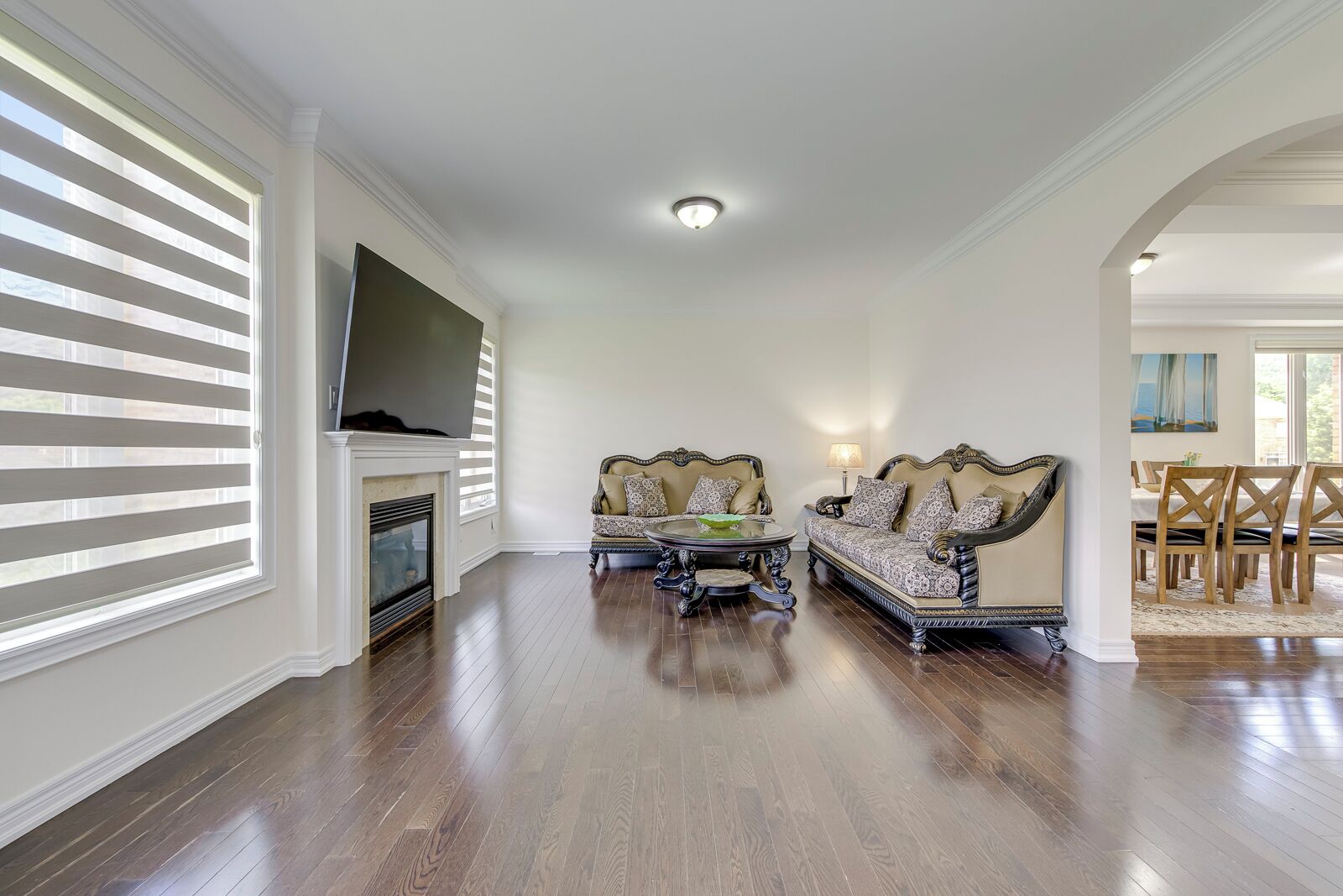 A spacious living room with hardwood flooring, two ornate sofas, a fireplace, and a television mounted on the wall.