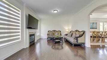A spacious living room with hardwood flooring, two ornate sofas, a fireplace, and a television mounted on the wall.