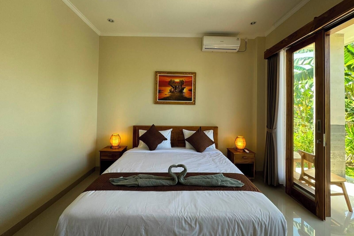 A hotel room with a bed, two bedside lamps, a painting on the wall, and a view of greenery through the sliding glass door.