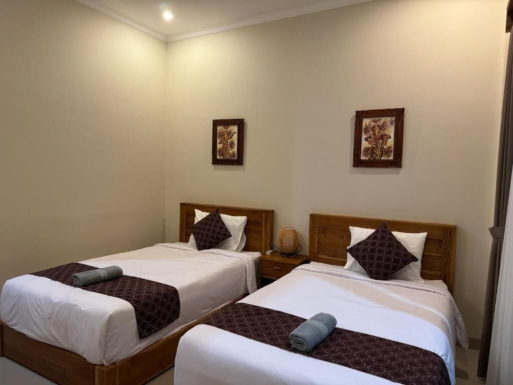Habitación de hotel con dos camas individuales, cada una con cabecero de madera, ropa de cama blanca y una manta marrón estampada. Hay dos mesitas de noche con lámparas, y las paredes están decoradas con cuadros.