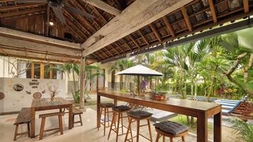 A spacious indoor area with a wooden ceiling, a bar area with stools, and a view of a pool with palm trees.