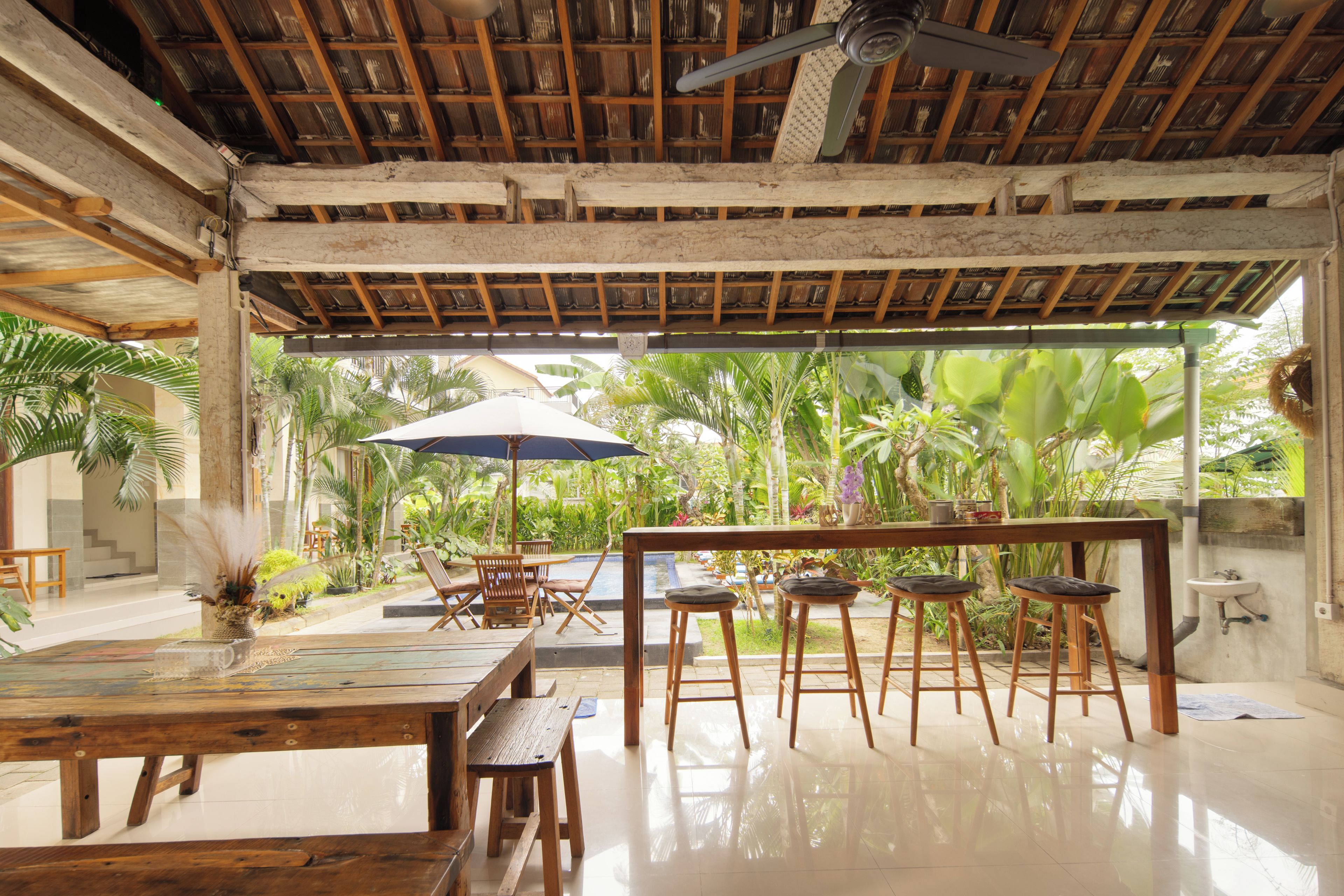 An indoor dining area with a wooden ceiling, a ceiling fan, a table with chairs, and a view of lush greenery outside.