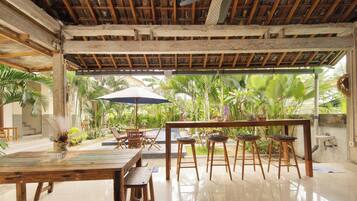 An indoor dining area with a wooden ceiling, a ceiling fan, a table with chairs, and a view of lush greenery outside.
