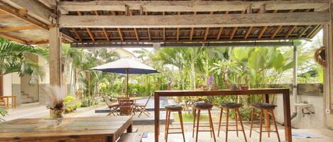 An indoor dining area with a wooden ceiling, a ceiling fan, a table with chairs, and a view of lush greenery outside.