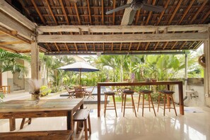 Un comedor interior con techo de madera, ventilador de techo, una mesa con sillas y vistas a la exuberante vegetación exterior.