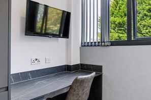 A modern room with a flat-screen TV mounted on the wall, a desk with a chair, and a window with blinds. - Brand New Spacious Studio in Beeston (Beeston)