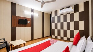 A hotel room with a bed, a desk, a chair, a television, and an air conditioning unit. - Hotel New Suryam (Ahmedabad)