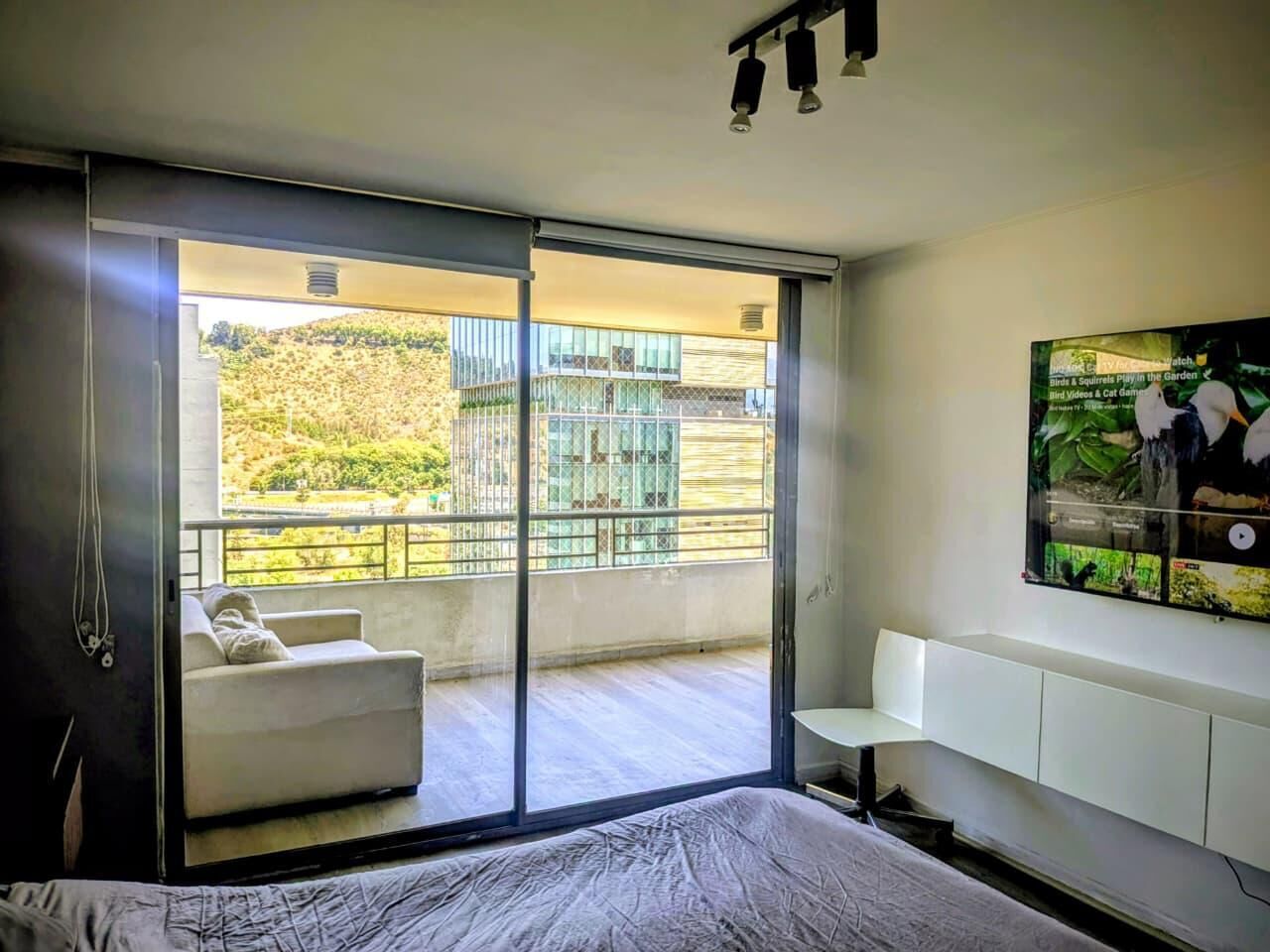 A bedroom with a sliding glass door leading to a balcony, a bed, a white sofa, and a mounted TV.