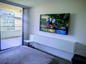 A hotel room with a bed, a TV mounted on the wall, a sliding glass door to the balcony, and a white bedside table. - Spectacular Golf Course: Newly Remodeled Apartment + Free Parking (Las Condes)