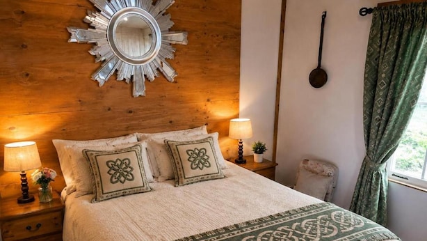 A cozy bedroom with a wooden headboard, a round mirror, two bedside lamps, a bed with a patterned throw, and a window with curtains. - Cottage Sleeps 4 With Parking and Sea Views (Lecamy)