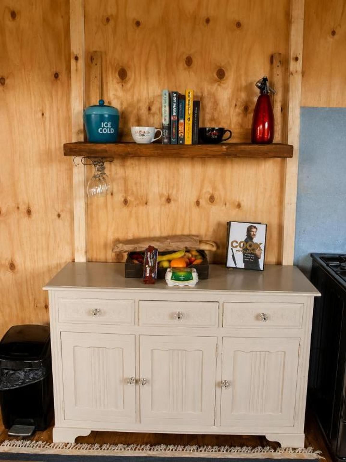En väggpanel av trä med en hylla där böcker, en mugg, en tekanna och en fotoram står. Under hyllan finns ett vitt skåp med lådor.