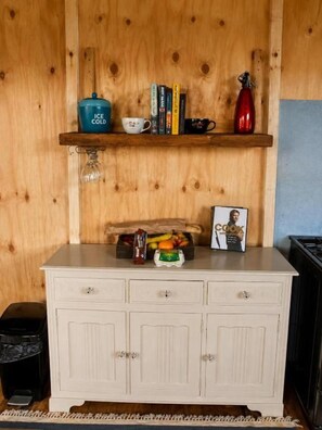 Pared de madera con un estante que sostiene libros, una taza, una tetera y un marco de fotos. Debajo del estante, un gabinete blanco con cajones.