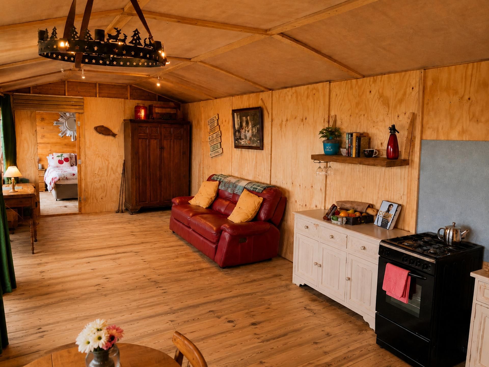 En mysig stuga med trägolv, en röd lädersoffa, ett matbord med en vas med blommor, ett köksområde med en spis och en hängande lampa.