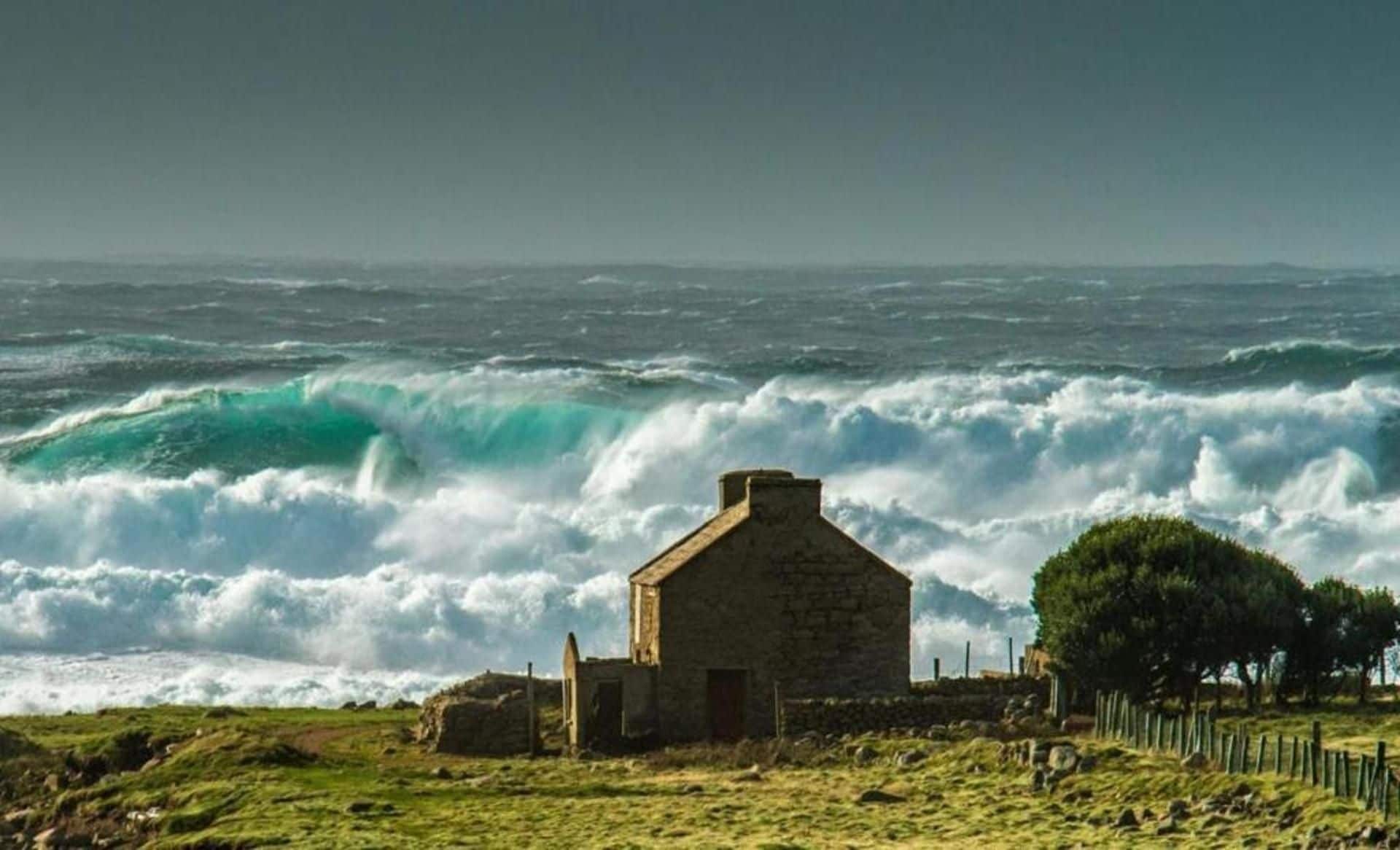 A coastal scene with a stone building, waves crashing, and a grassy area with trees.