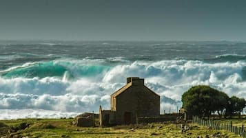 A coastal scene with a stone building, waves crashing, and a grassy area with trees.