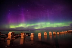 Escena nocturna con la aurora boreal sobre una superficie acuática tranquila y una fila de postes de madera.