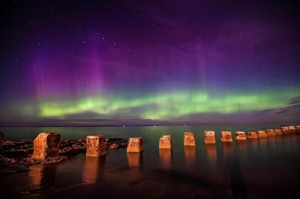 A night scene with the Northern Lights over a calm body of water and a row of wooden posts. - Seaside Retreat Cottage in Carrowmenagh (Lecamy)