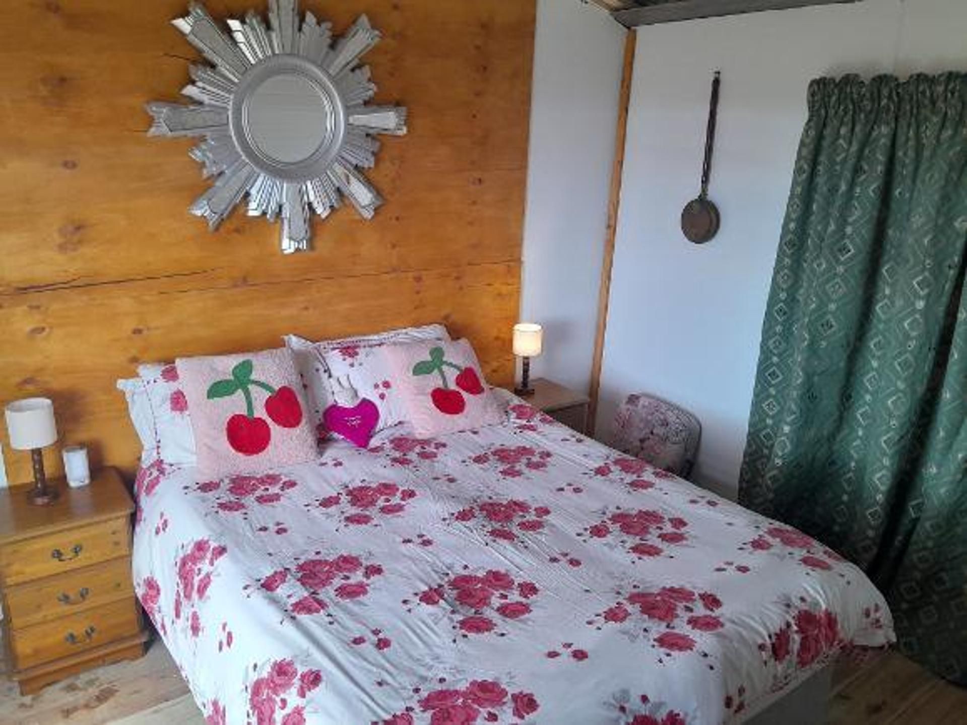 A bedroom with a wooden headboard, a large mirror, a bed with floral bedding, a bedside table with a lamp, and a green patterned curtain.