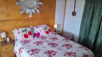 A bedroom with a wooden headboard, a large mirror, a bed with floral bedding, a bedside table with a lamp, and a green patterned curtain.