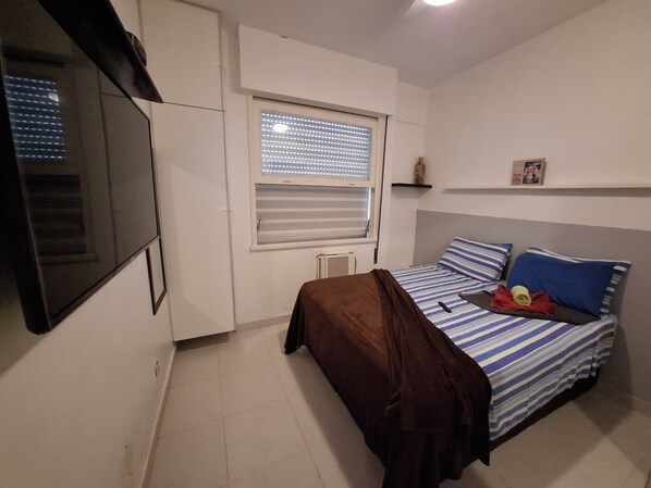 Un dormitorio con cama, ventana con persianas, radiador y televisor montado en la pared.