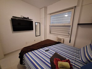 Un dormitorio con cama, televisor montado en la pared y ventana con persianas.