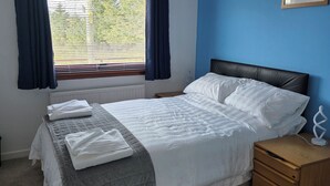A neatly made bed with white linens and a gray quilt, a wooden nightstand with a lamp, and a window with blue curtains. - 7 Creag Ruadh (Isle of Lewis)