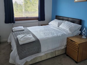 Una cama bien tendida con sábanas blancas y colcha gris, una mesita de noche de madera con una lámpara, y una ventana con cortinas azules.