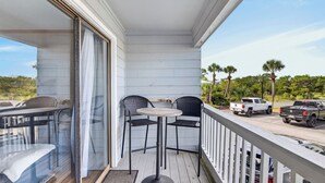 En balkon med to stole og et bord, med udsigt over en parkeringsplads med køretøjer og palmer.