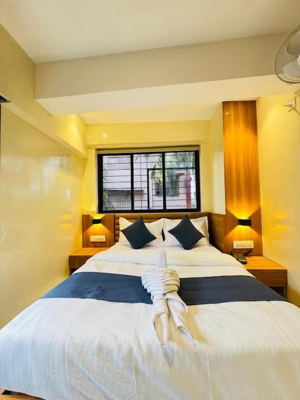 A hotel room with a bed, two bedside lamps, a window with blinds, and a fan on the ceiling. - Hotel Comfort Castle (Mumbai)