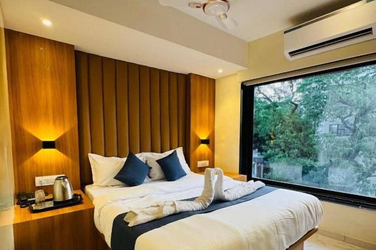 A modern hotel room with a large bed, wooden panel wall, bedside lamps, and a view of greenery through a large window.