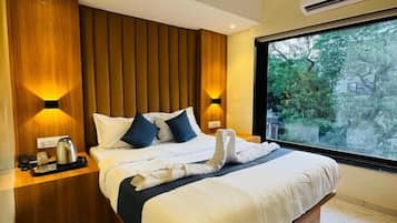 A modern hotel room with a large bed, wooden panel wall, bedside lamps, and a view of greenery through a large window.