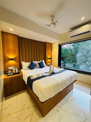 A modern hotel room with a large bed, wooden panel wall, bedside lamps, and a view of greenery through a large window. - Hotel Comfort Castle (Mumbai)