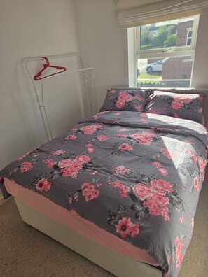 A bed with a floral bedspread, a red hanger on a metal rack, and a window with a view of a car and greenery. - Shirebrook 2-bed Townhouse, With Free Parking, Ideal for Contractors and Families (Mansfield)