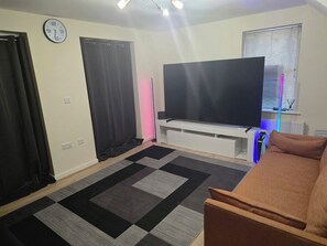 A living room with a flat-screen TV, a sofa, a patterned rug, and a window with blinds. - Shirebrook 2-bed Townhouse, With Free Parking, Ideal for Contractors and Families (Mansfield)
