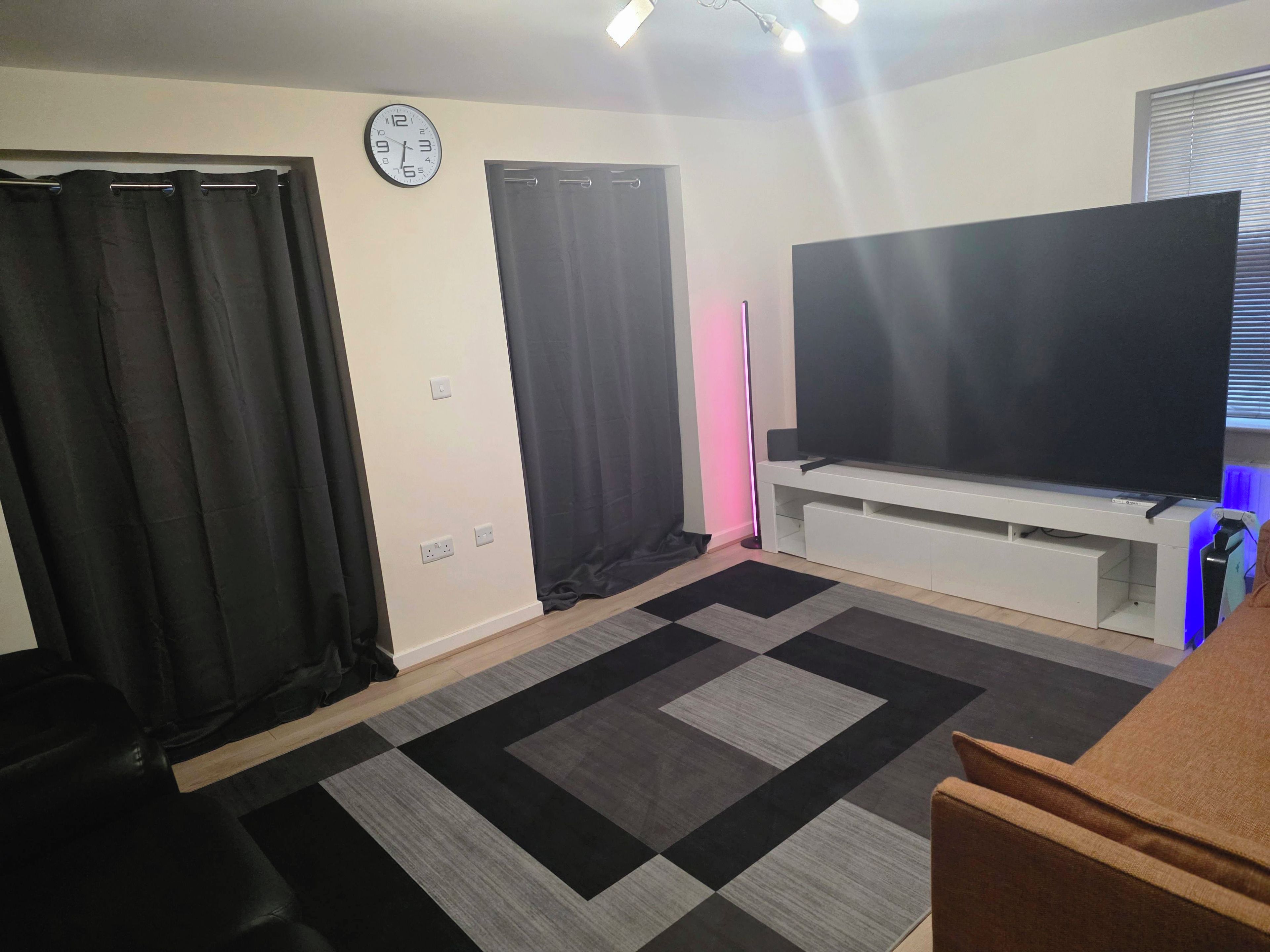 A room with a flat-screen TV on a stand, a clock on the wall, and a patterned rug.