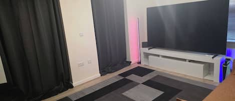 A room with a flat-screen TV on a stand, a clock on the wall, and a patterned rug.