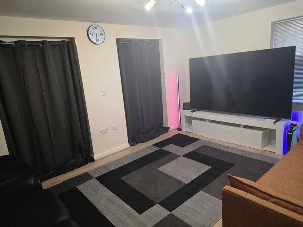 A room with a flat-screen TV on a stand, a clock on the wall, and a patterned rug.