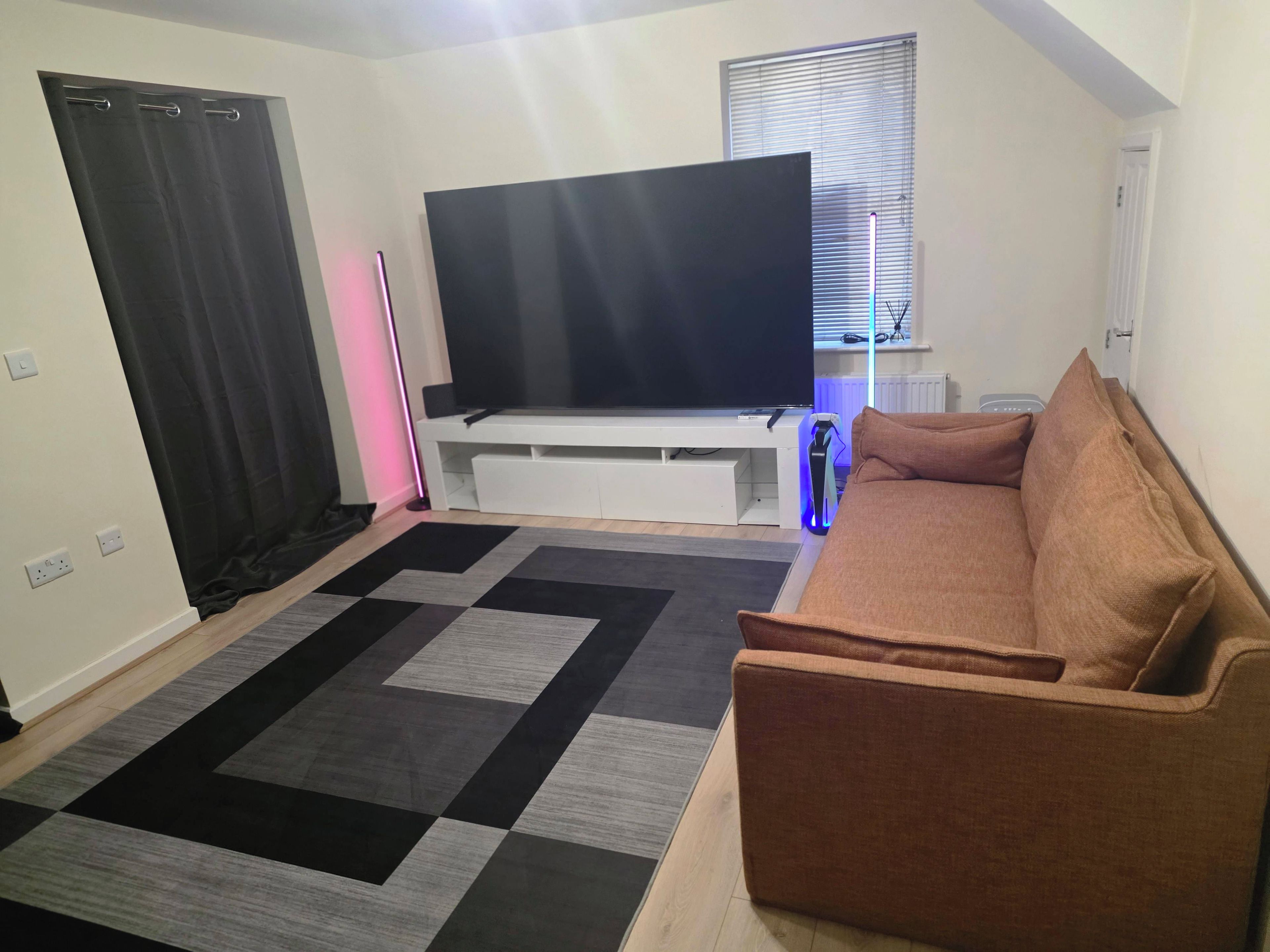 Ein Wohnzimmer mit einem Flachbildfernseher auf einem weißen Schrank, einem braunen Sofa und einem gemusterten Teppich.