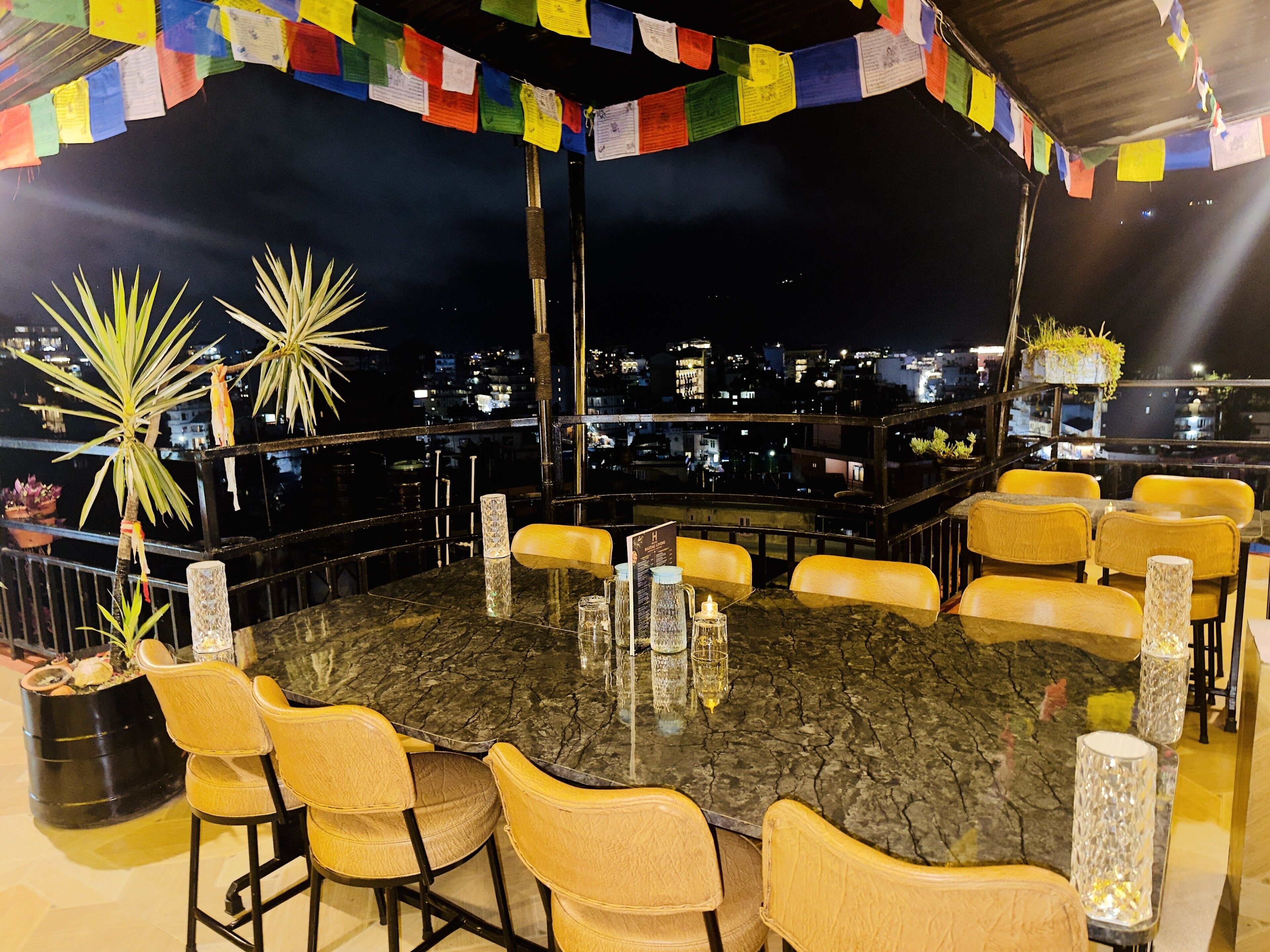 A rooftop dining area with a round table surrounded by chairs, a view of the cityscape, and colorful prayer flags.