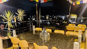 Ein Essbereich auf dem Dach mit einem runden Tisch, umgeben von Stühlen, mit Blick auf die Stadtlandschaft und bunten Gebetsfahnen.
