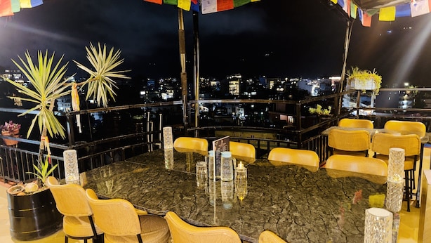 Un área de comedor en la azotea con una mesa redonda rodeada de sillas, vista del paisaje urbano y banderas de oración coloridas.