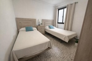 A room with two single beds, a nightstand with a lamp, a window with curtains, and a speckled floor. - Apartamento Canarias (Los Cristianos)