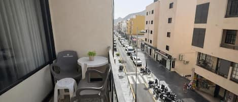 A balcony with a table and chairs, a potted plant, and a view of a street with parked cars and buildings.