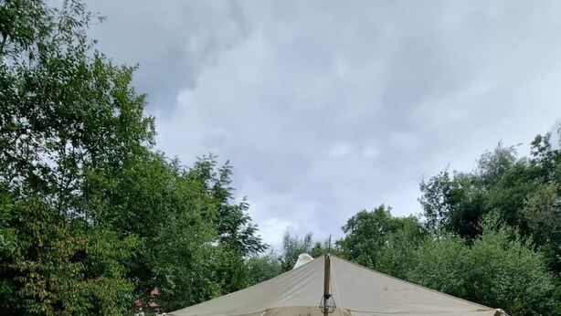 Ein Zelt mit Bett, Tisch und Stühlen, auf einer Wiese vor einer Baumkulisse.