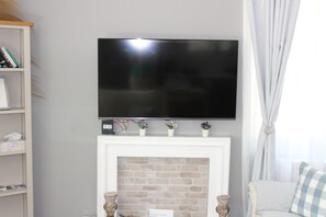 A living room with a flat-screen TV mounted above a fireplace, a bookshelf to the left, and a window with curtains to the right. -  1-bedroom bungalow Limassol Countryside Tranquil Getaway Shared Pool Near Beach (Limassol)