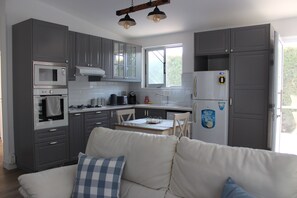 A compact kitchen with modern appliances, a dining area with a table and chairs, and a sofa with a checkered pillow. -  1-bedroom bungalow Limassol Countryside Tranquil Getaway Shared Pool Near Beach (Limassol)